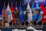 A man in a Army dress uniform stands and speaks on stage with multiple flags behind him in a large ballroom.
