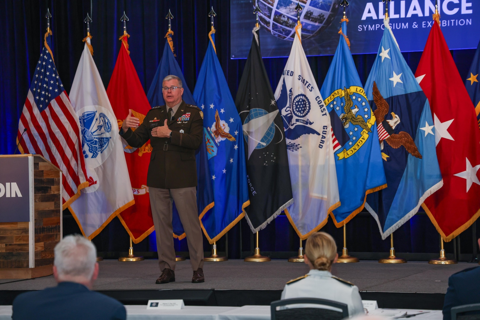 A man in a Army dress uniform stands and speaks on stage with multiple flags behind him in a large ballroom.