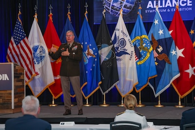 A man in a Army dress uniform stands and speaks on stage with multiple flags behind him in a large ballroom.