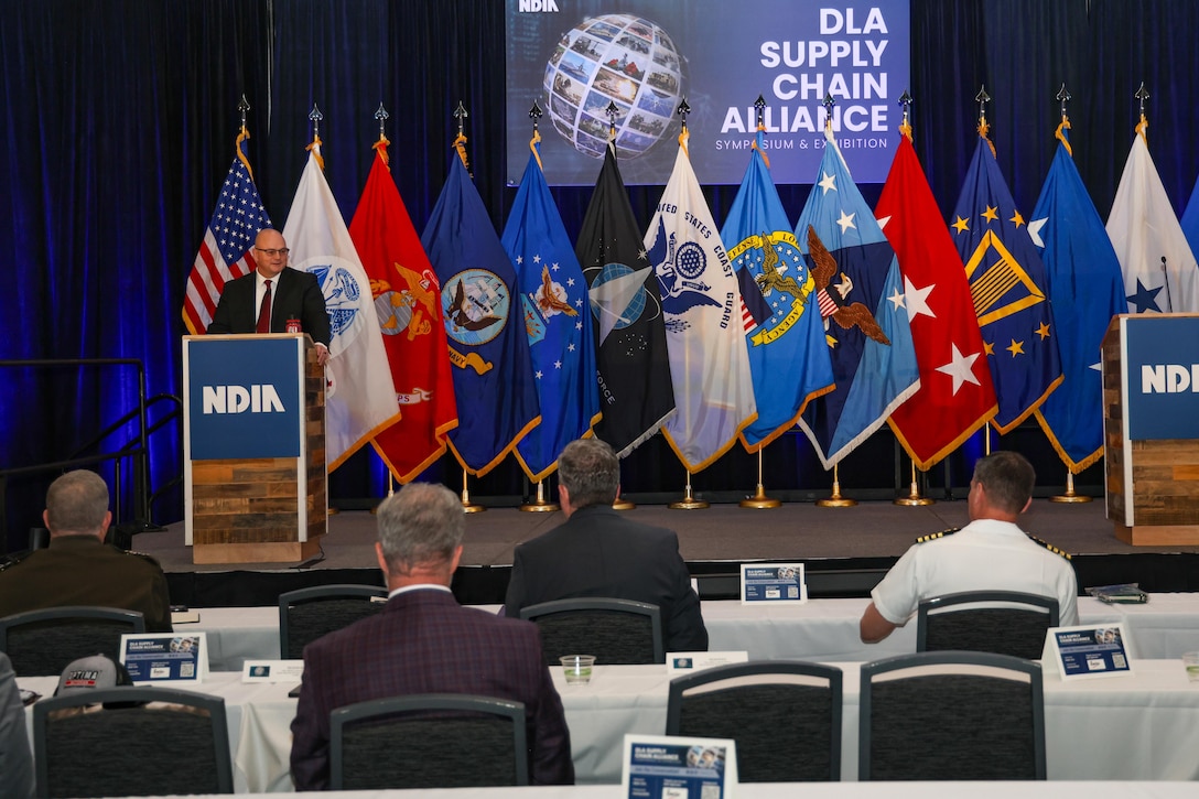 A man in a business suit speaks at a podium on a large stage with multiple flags behind in a large ballroom.