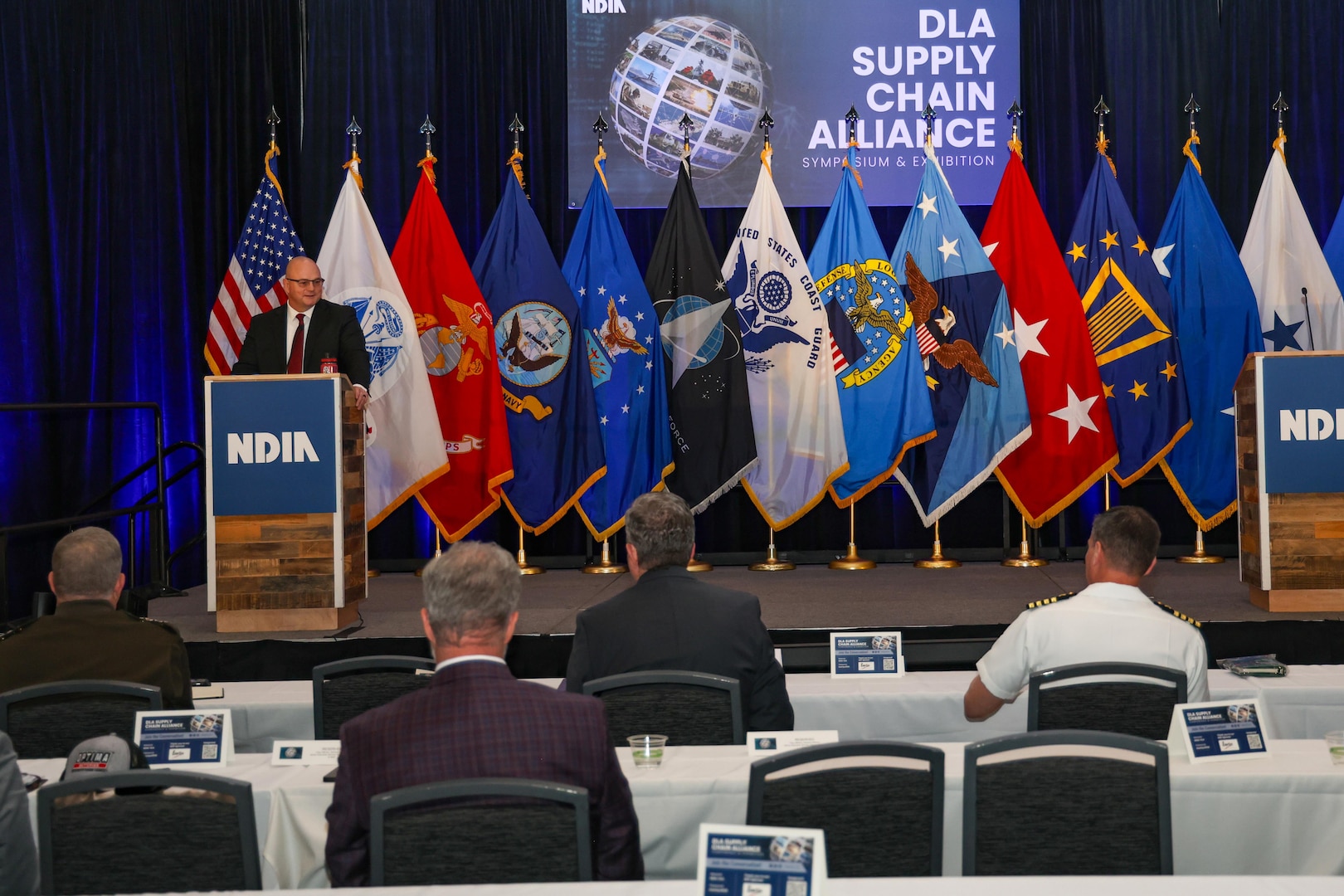 A man in a business suit speaks at a podium on a large stage with multiple flags behind in a large ballroom.