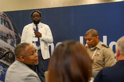 A main in a striped suit speaks on stage in a exhibit hall.
