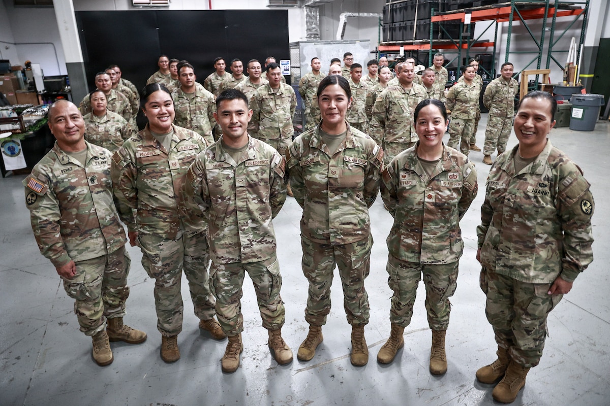 "Brig. Gen. Karin Watson, Command Sgt. Maj. Duane Perez, and Maj. Audi Taitano stand in the front row, smiling with a large group of uniformed service members for a celebratory photo. The squadron is assembled inside a warehouse-like space.