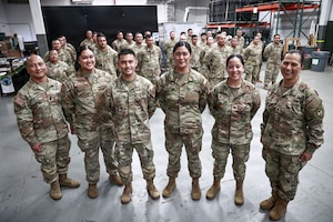 "Brig. Gen. Karin Watson, Command Sgt. Maj. Duane Perez, and Maj. Audi Taitano stand in the front row, smiling with a large group of uniformed service members for a celebratory photo. The squadron is assembled inside a warehouse-like space.