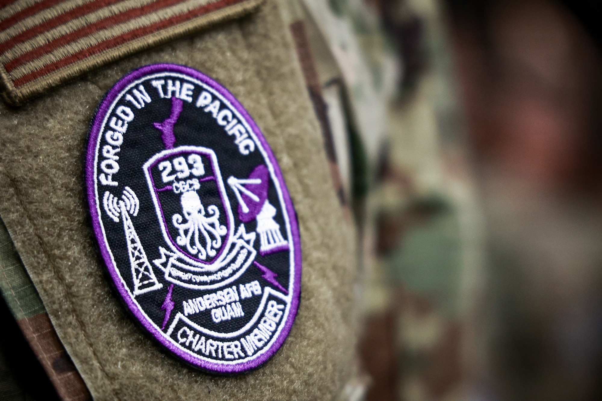 A close-up photo focuses on the circular, purple-bordered patch of the 293rd Combat Communications Squadron on the sleeve of a uniform. Text on the patch reads, 'FORGED IN THE PACIFIC,' 'ANDERSEN AFB GUAM,' and 'CHARTER MEMBER,' with an octopus emblem in the center.