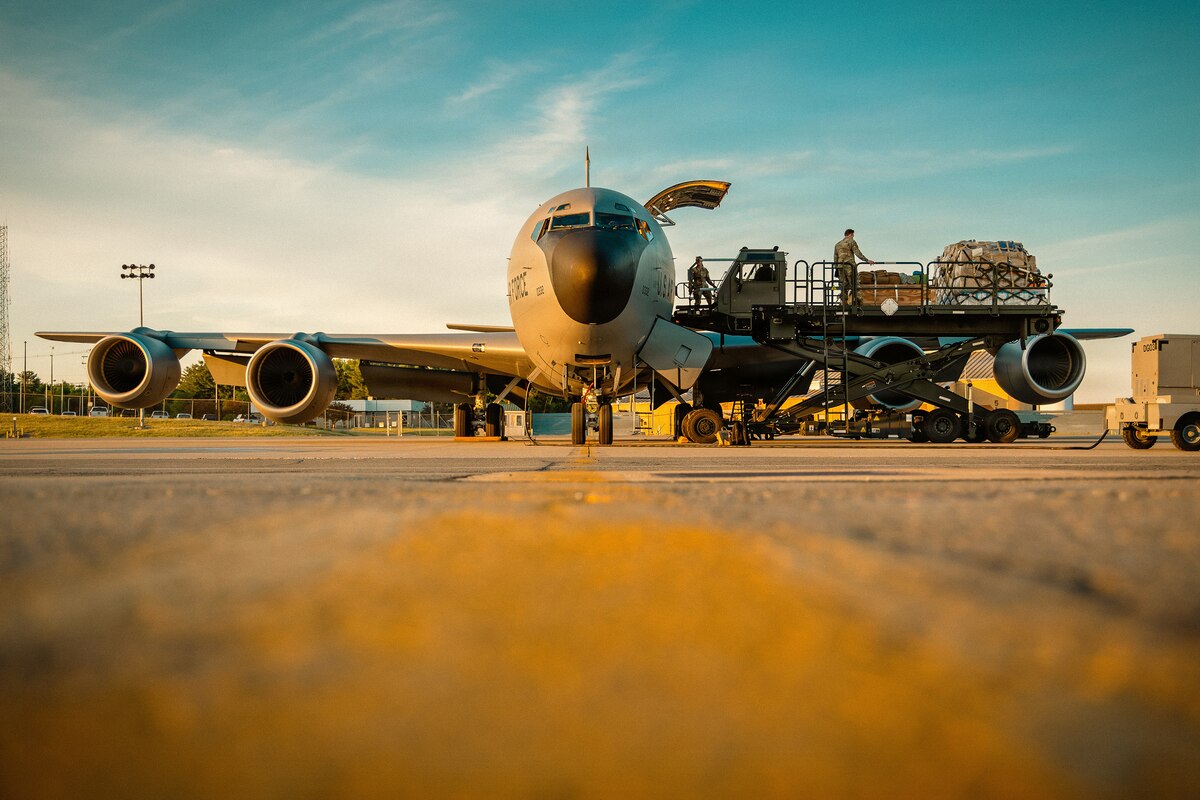 A large military aircraft sits on a flight line with the door open as people in camouflage military uniforms load cargo.