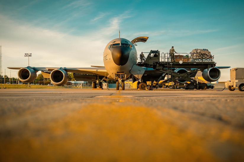 A large military aircraft sits on a flight line with the door open as people in camouflage military uniforms load cargo.