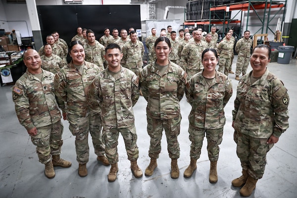 "Brig. Gen. Karin Watson, Command Sgt. Maj. Duane Perez, and Maj. Audi Taitano stand in the front row, smiling with a large group of uniformed service members for a celebratory photo. The squadron is assembled inside a warehouse-like space.