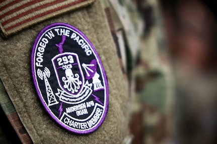 A close-up photo focuses on the circular, purple-bordered patch of the 293rd Combat Communications Squadron on the sleeve of a uniform. Text on the patch reads, 'FORGED IN THE PACIFIC,' 'ANDERSEN AFB GUAM,' and 'CHARTER MEMBER,' with an octopus emblem in the center.