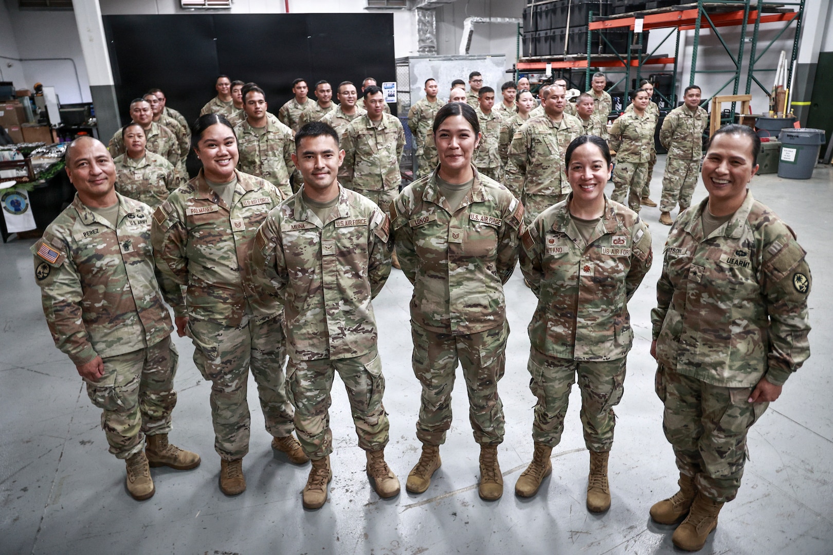 "Brig. Gen. Karin Watson, Command Sgt. Maj. Duane Perez, and Maj. Audi Taitano stand in the front row, smiling with a large group of uniformed service members for a celebratory photo. The squadron is assembled inside a warehouse-like space.