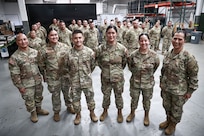 "Brig. Gen. Karin Watson, Command Sgt. Maj. Duane Perez, and Maj. Audi Taitano stand in the front row, smiling with a large group of uniformed service members for a celebratory photo. The squadron is assembled inside a warehouse-like space.