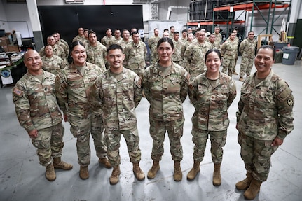 "Brig. Gen. Karin Watson, Command Sgt. Maj. Duane Perez, and Maj. Audi Taitano stand in the front row, smiling with a large group of uniformed service members for a celebratory photo. The squadron is assembled inside a warehouse-like space.