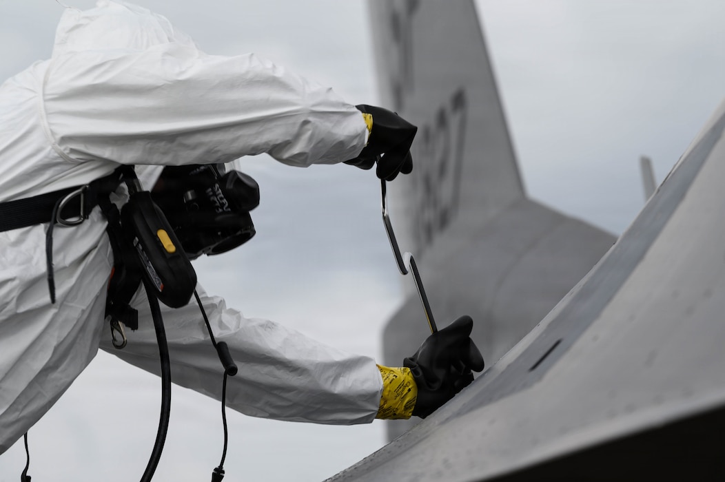 A man in a hazmat suit unbolts a panel on an F-16 Fighting Falcon fighter jet.