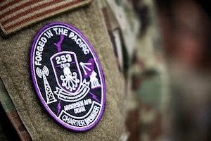 A close-up view of a sleeve on a U.S. military uniform. There is a round patch on the sleeve with a black background and purple accents. Text along the top reads: Forged in the pacific. Text along the bottom reads: Charter member.