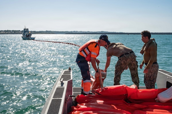 Sailors and local nationals drop orange inflatable booms into the water off a boat.