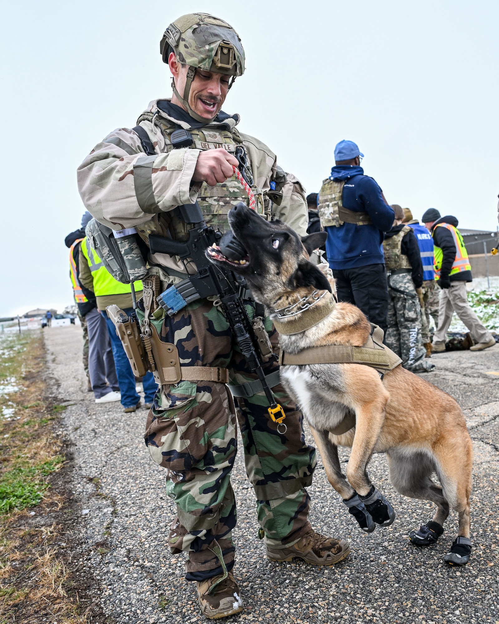 Airman in camouflage gear using a dog toy on a rope to play tug of war with a Belgian Malinois wearing protective boots on a leash outdoors.
