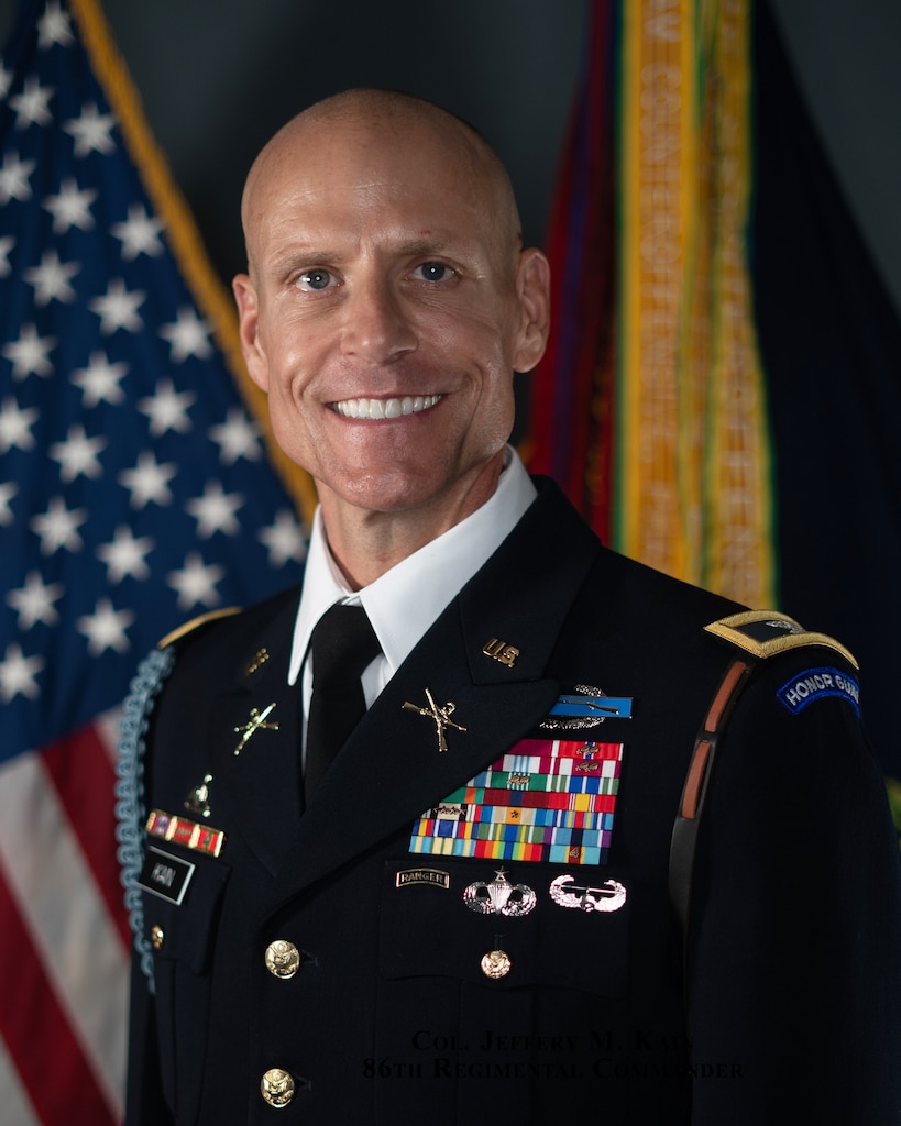 Posed portrait photo of U.S. Army colonel in dark blue service dress uniform with flags in the background.