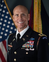 Posed portrait photo of U.S. Army colonel in dark blue service dress uniform with flags in the background.