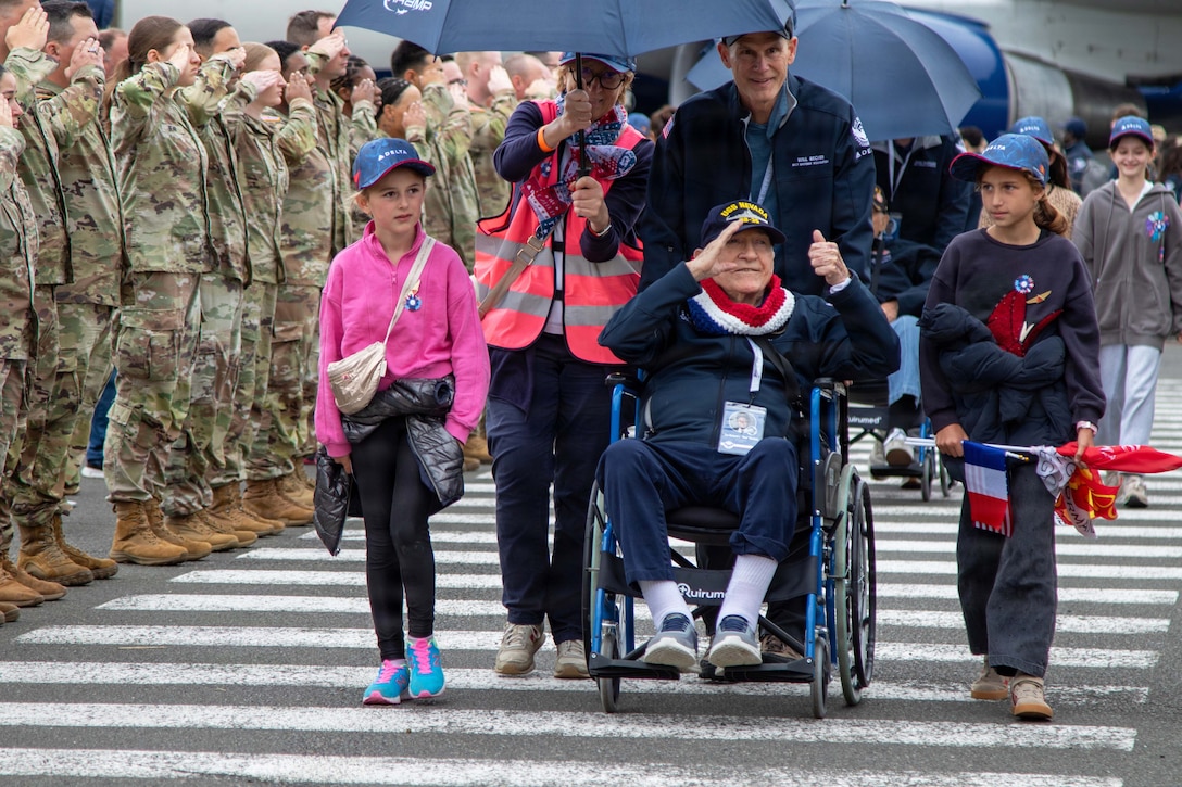 Loved ones push veterans in a wheelchair under an umbrella in a crosswalk as service members salute while standing in formation.