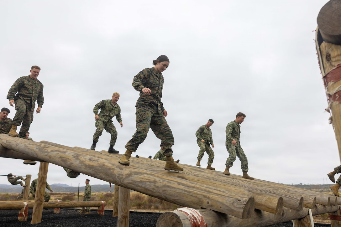 Navy midshipmen walk down log obstacles against a gray sky.