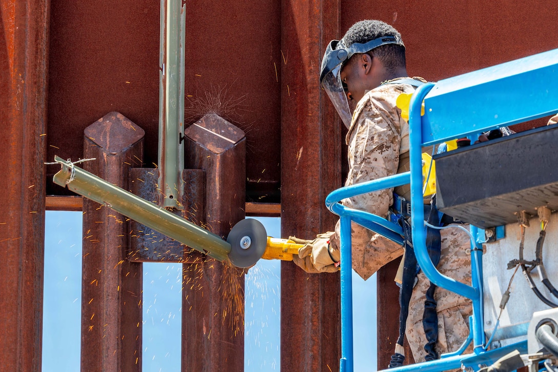 Sparks fly as a Marine in a blue lift wearing protective gear cuts through metal attached to a large fence.