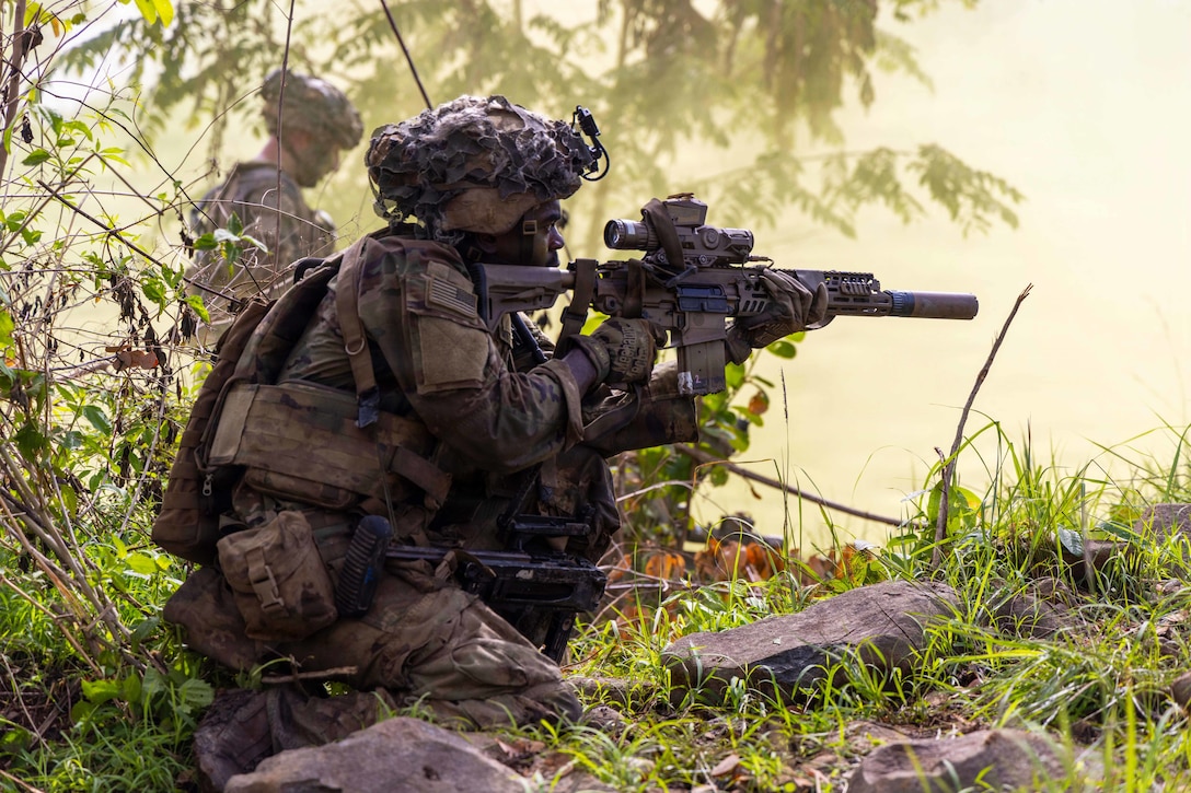 A soldier in camouflage holds a rifle while kneeling on the ground in a wooded area. Another soldier is in the background.