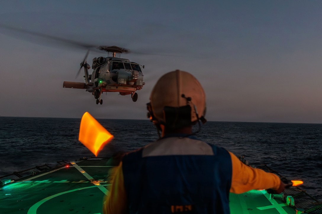 A sailor in a yellow helmet uses yellow light wands to help a pilot land a helicopter on the flight deck of a ship that is lit with green lights.