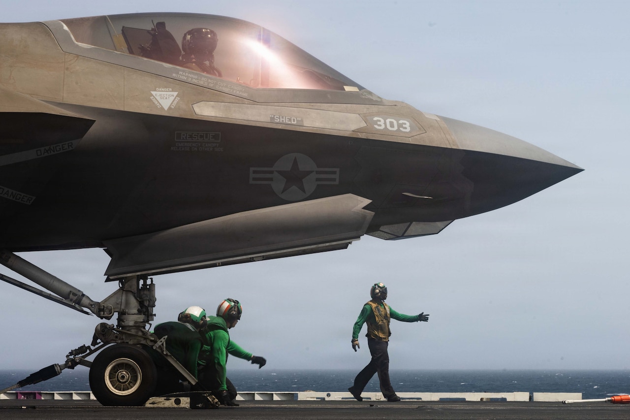 Sailors in green shirts and helmets stand on a flight deck near the nose of a fighter jet, with the horizon in the background.