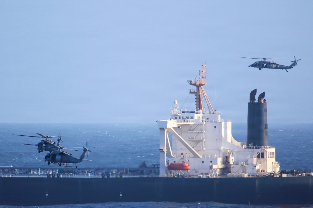 Military helicopters hover over a vessel, with the horizon in the background.