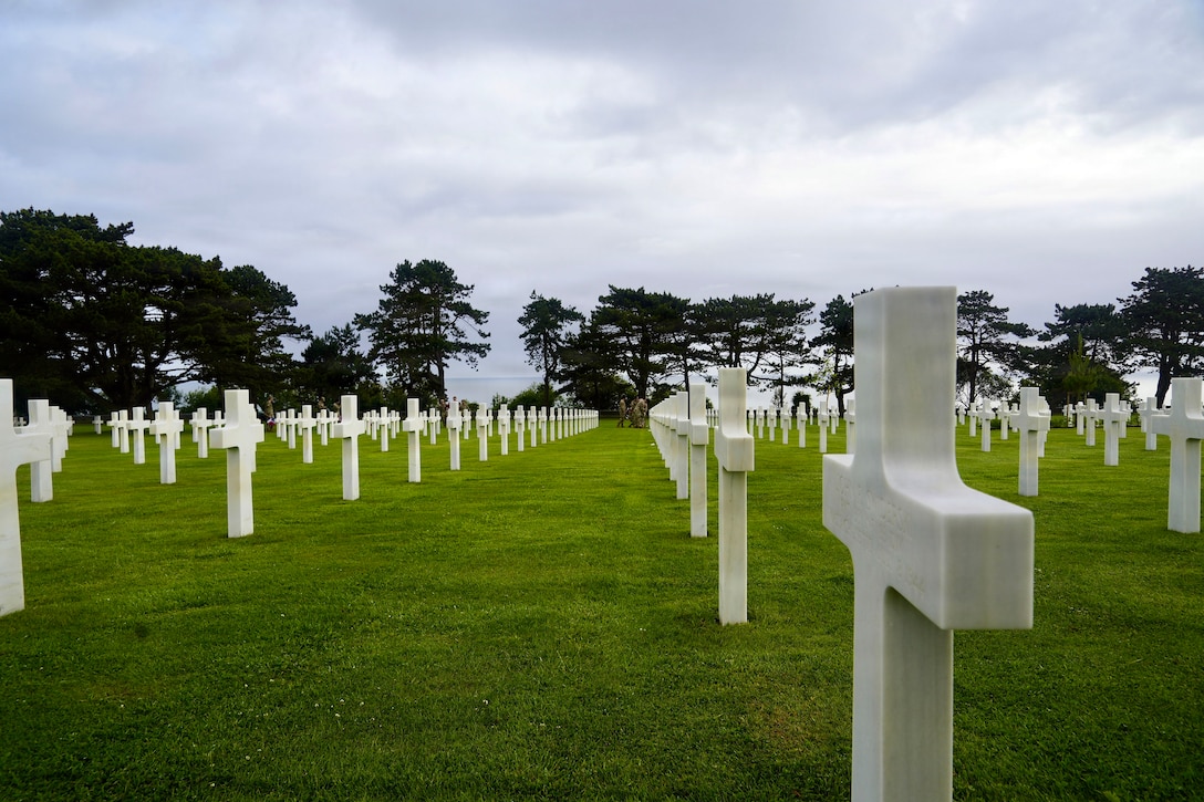 Rows of white headstones, mostly crosses, line a green field with trees in the distrance.