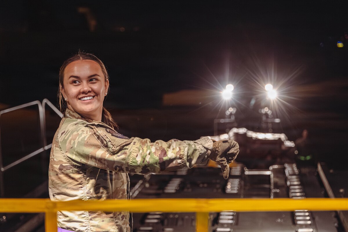 A woman in a camouflage military uniform smiles and looks back while giving a thumbs down during the evening hours on a flight line. Vehicle headlights shine in the distance.