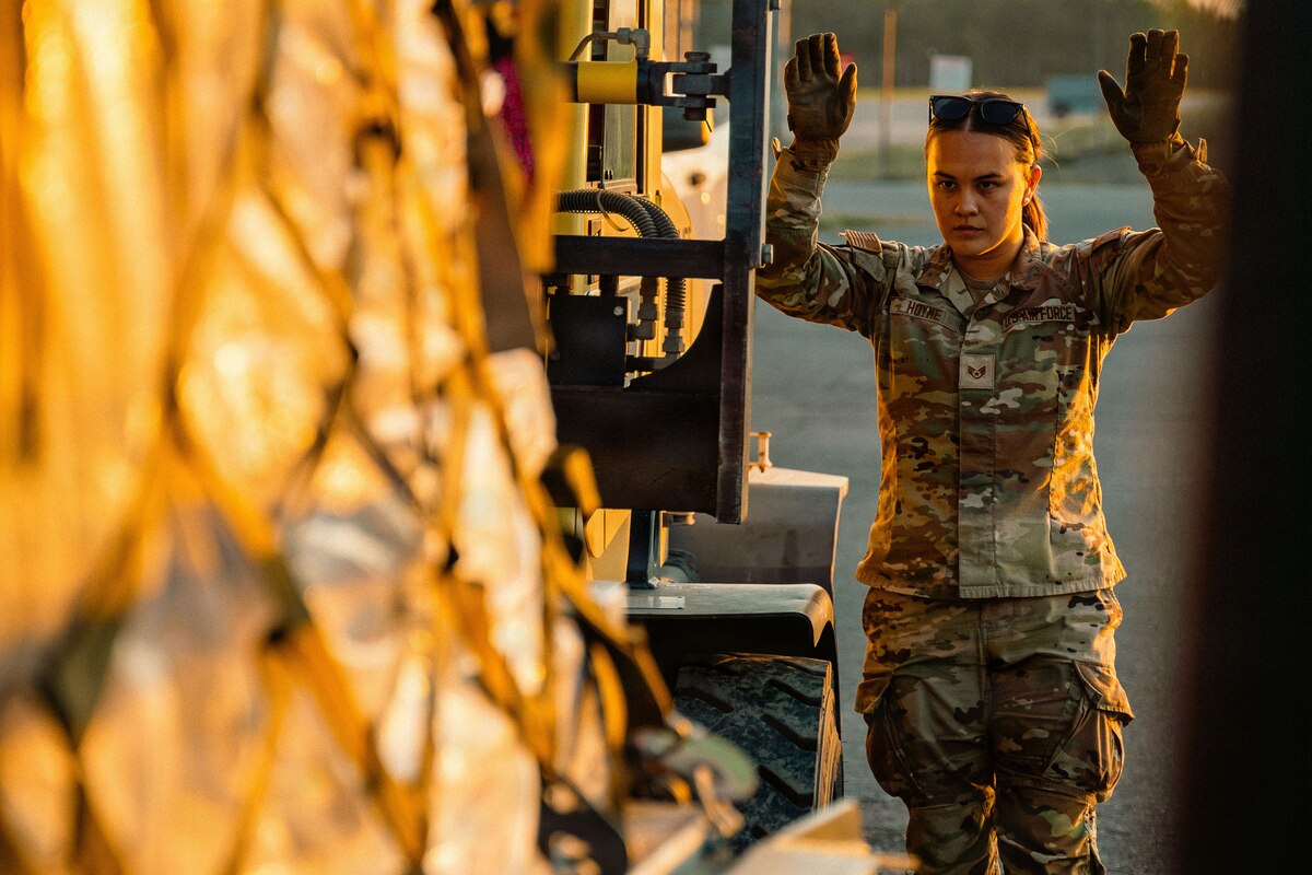 A woman in a camouflage military uniform directs a military loading machine carrying a pallet of supplies.