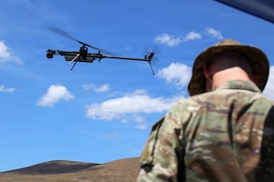A drone flies in a blue sky with clouds as a rear-facing person in a camouflage military uniform looks down to pilot it; hills are in the distance.