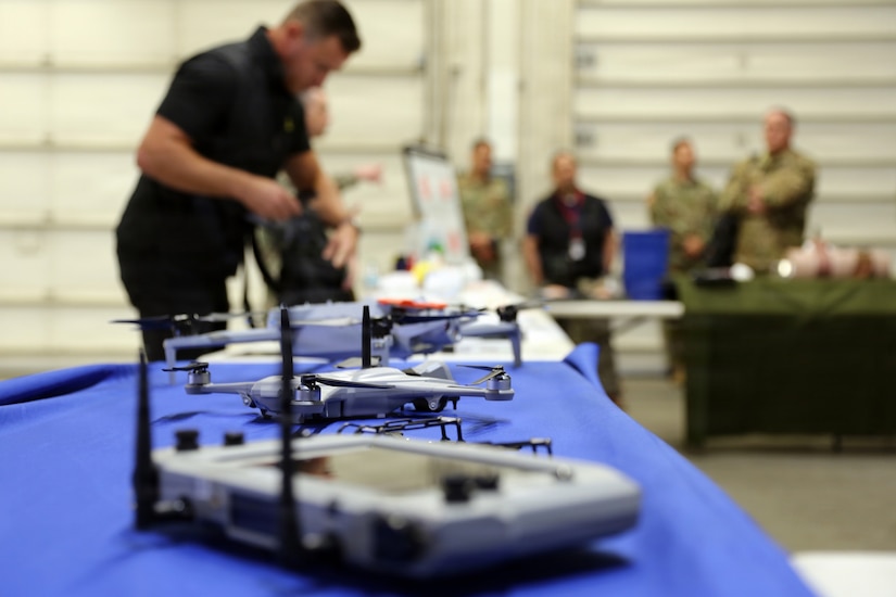 A close-up of several drones and remote controls sitting on a table; people in camouflage military uniforms and others in casual attire stand in the background.