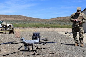 A drone sits on rocky terrain as a man in a camouflage military uniform looks down to pilot it; people in similar attire are in the background, and hills are in the distance.