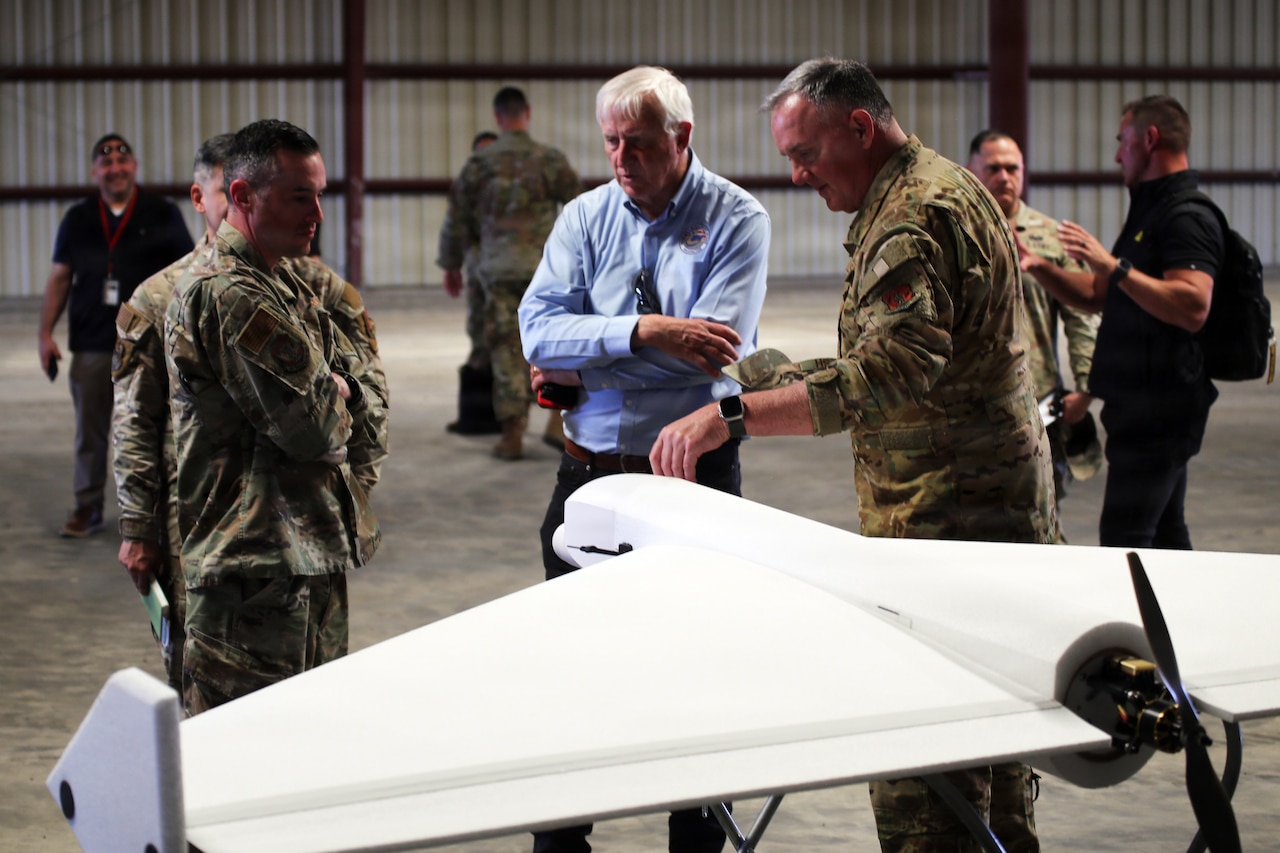 Nearly a dozen people, some in camouflage military uniforms and others in casual attire, stand and talk in a hangar; a white drone sits on the floor in the foreground.