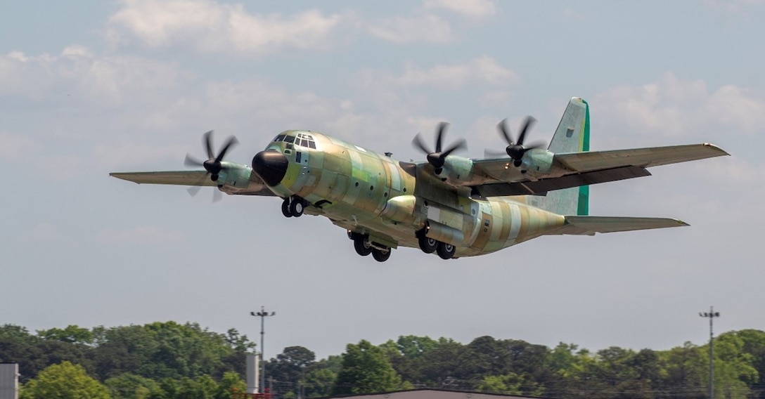 A green or unpainted C-130J rises above the tree line after takeoff.