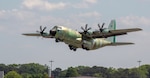 A green or unpainted C-130J rises above the tree line after takeoff.