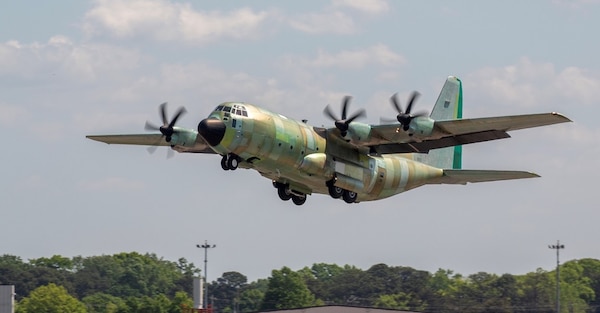 A green or unpainted C-130J rises above the tree line after takeoff.