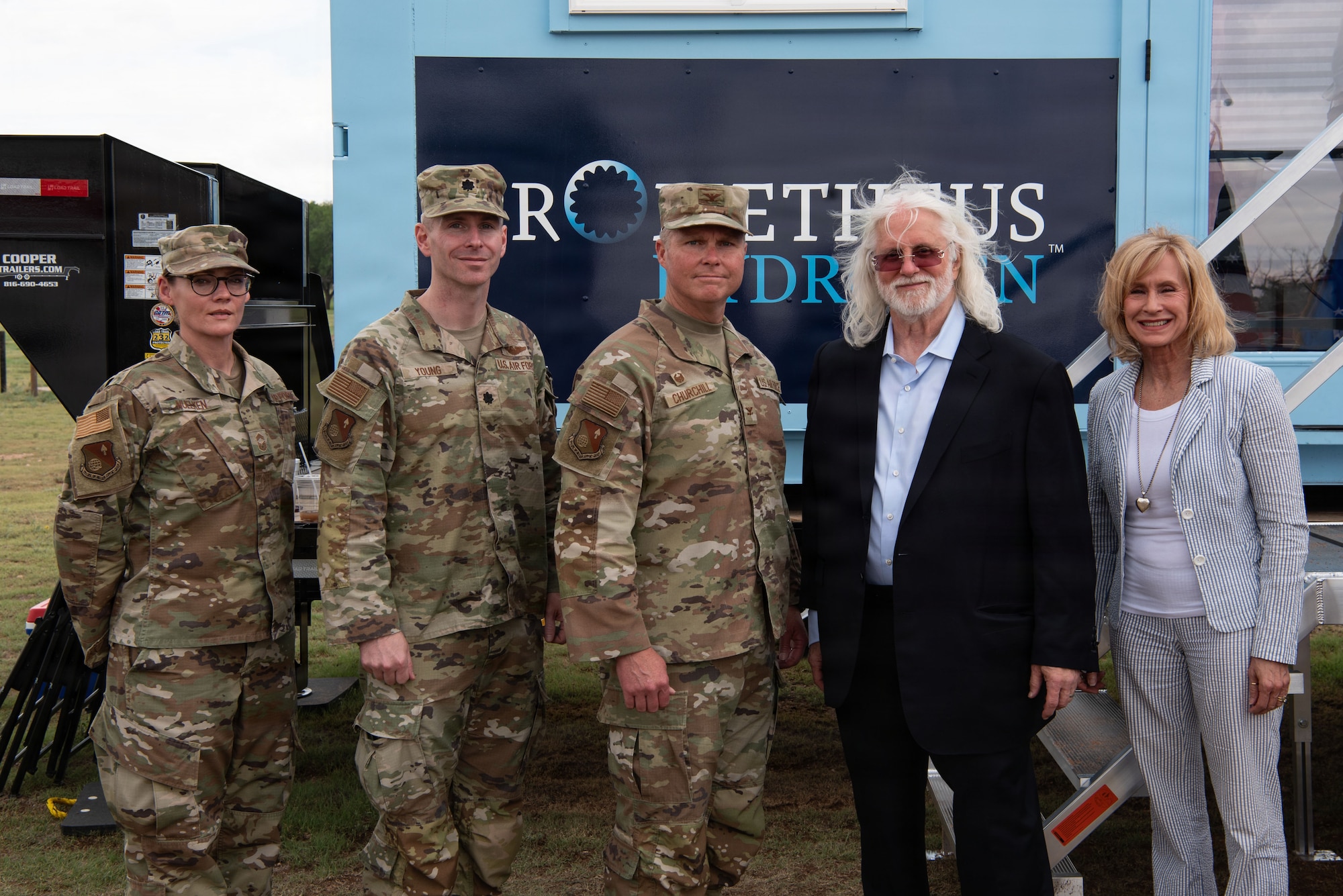 a group of give people, three in uniform, two in suits, stand together in front of a blue box