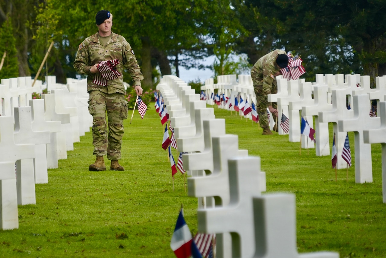 Two soldiers work to place small U.S. flags at white cross-shaped headstones, many already adorned with U.S. and French flags.
