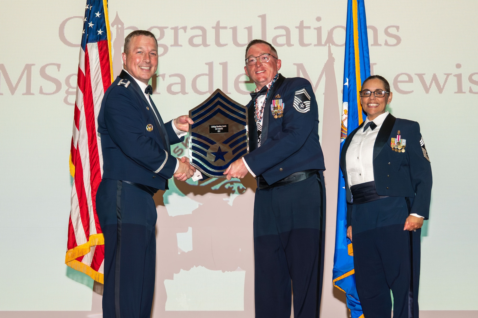 U.S. Air Force Senior Master Sgt. Bradley Lewis, 341st Munitions Squadron senior enlisted leader, center, receives a plaque from Col. Daniel Voorhies, 341st Missile Wing commander, in recognition of his advancement to the pinnacle of the enlisted rank structure, during the 2026 Chief Master Sgt. recognition ceremony at Malmstrom Air Force Base, Montana, May 29, 2026.