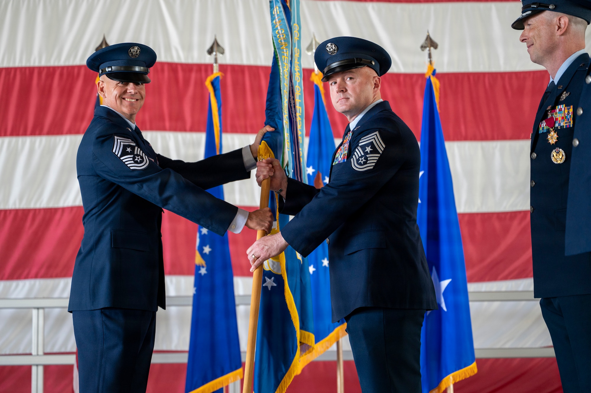 U.S. Air Force Chief Master Sgt. William Ford passes the 49th Wing guidon to Chief Master Sgt. Scott Piper during a change of command ceremony at Holloman Air Force Base, New Mexico, June 4, 2026. Ford served as the 49th Wing command chief for two years, leading and developing the Fightin’ 49ers to become skilled Airmen. (U.S. Air Force photo by Senior Airman Michelle Ferrari)