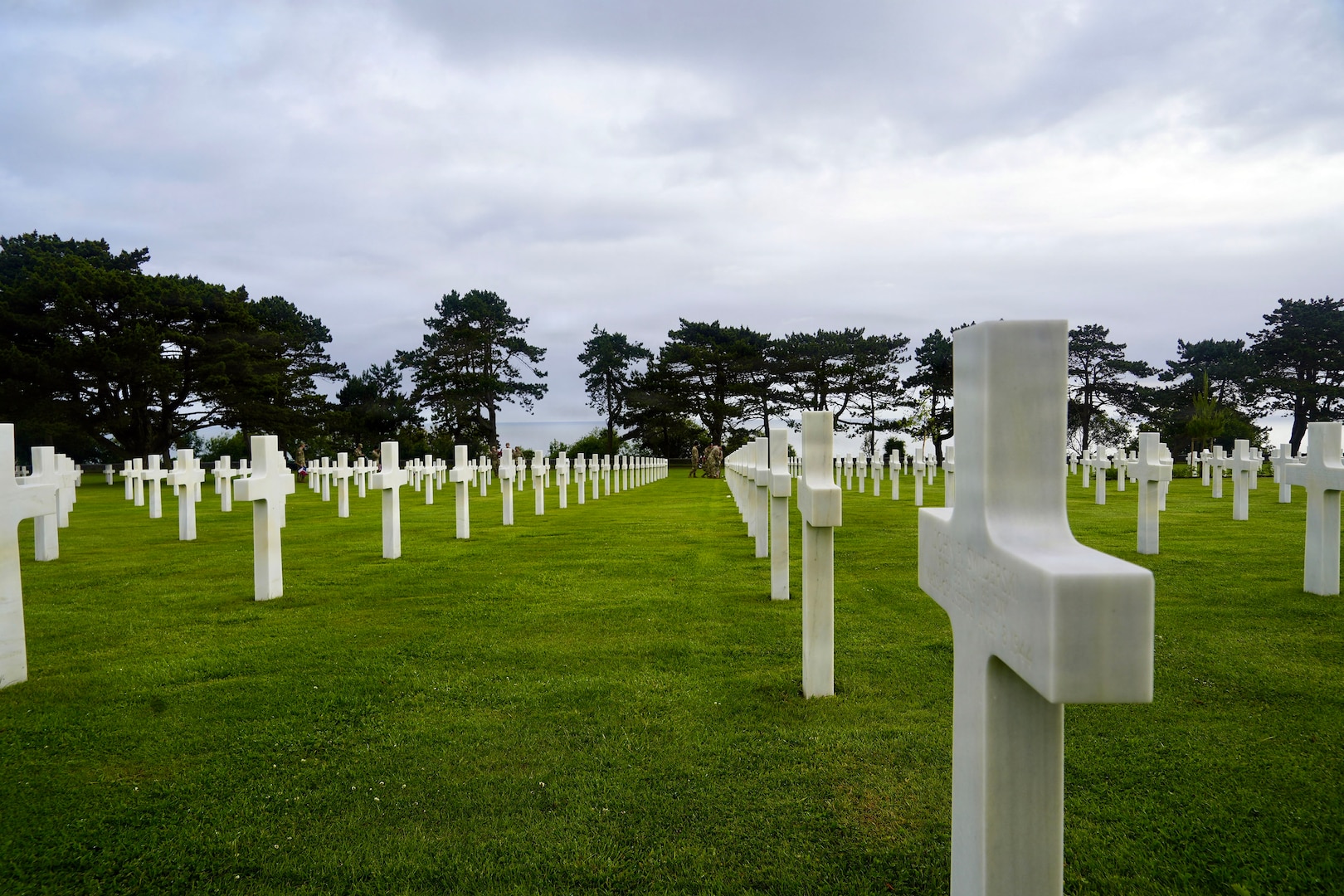 Rows of white headstones, mostly crosses, line a green field with trees in the distrance.