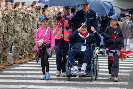Loved ones push veterans in a wheelchair under an umbrella in a crosswalk as service members salute while standing in formation.