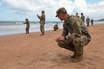 Soldiers using small handheld tools walk or kneel on a sandy beach during the day.