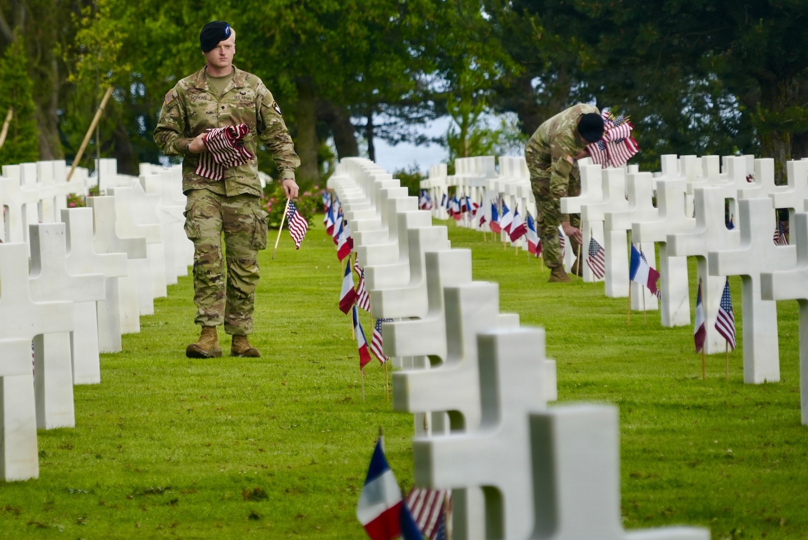 Two soldiers work to place small U.S. flags at white cross-shaped headstones, many already adorned with U.S. and French flags.