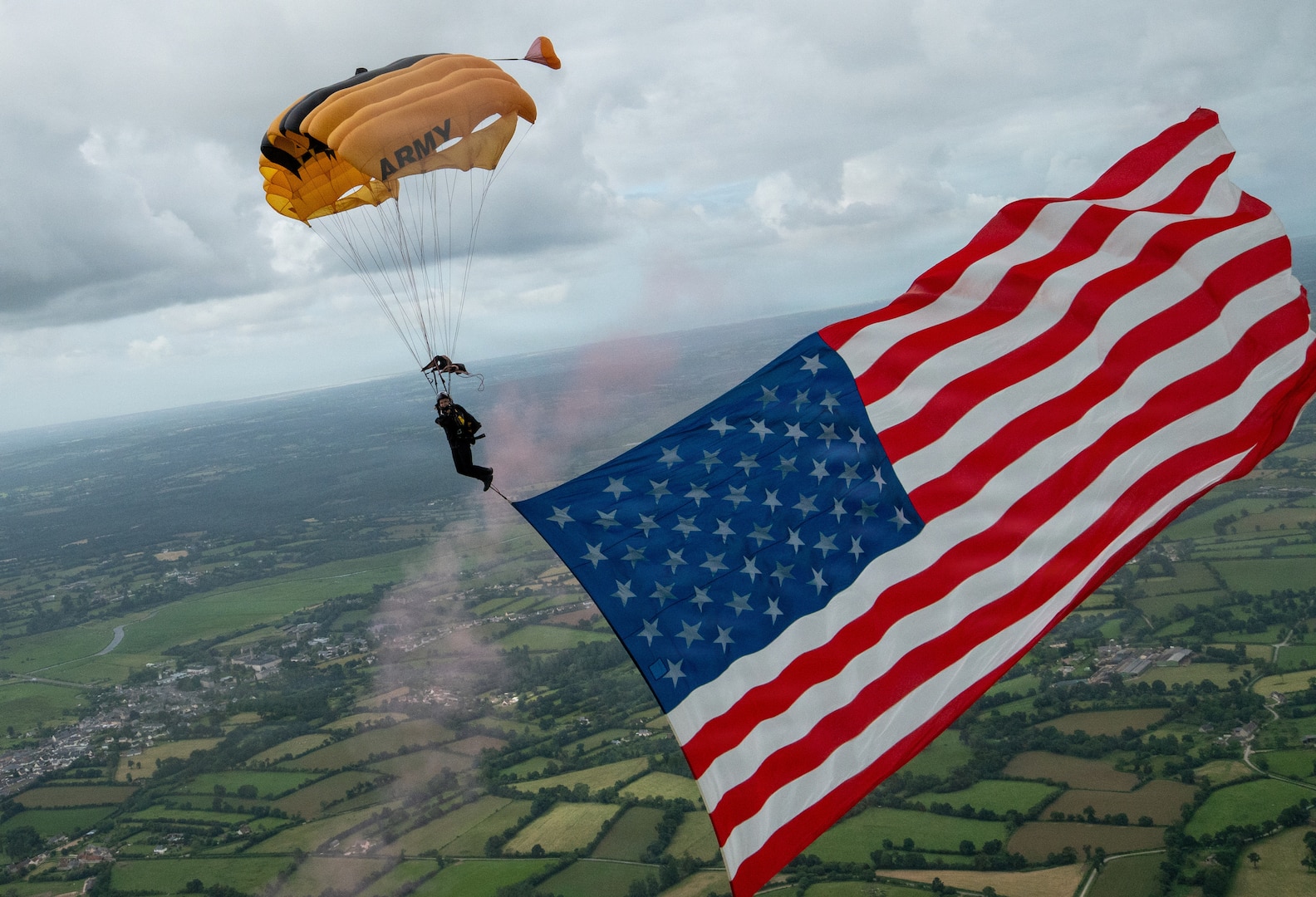 A soldier with a yellow parachute descends of green terrain, a U.S. flag affixed to their feet waving in the breeze.