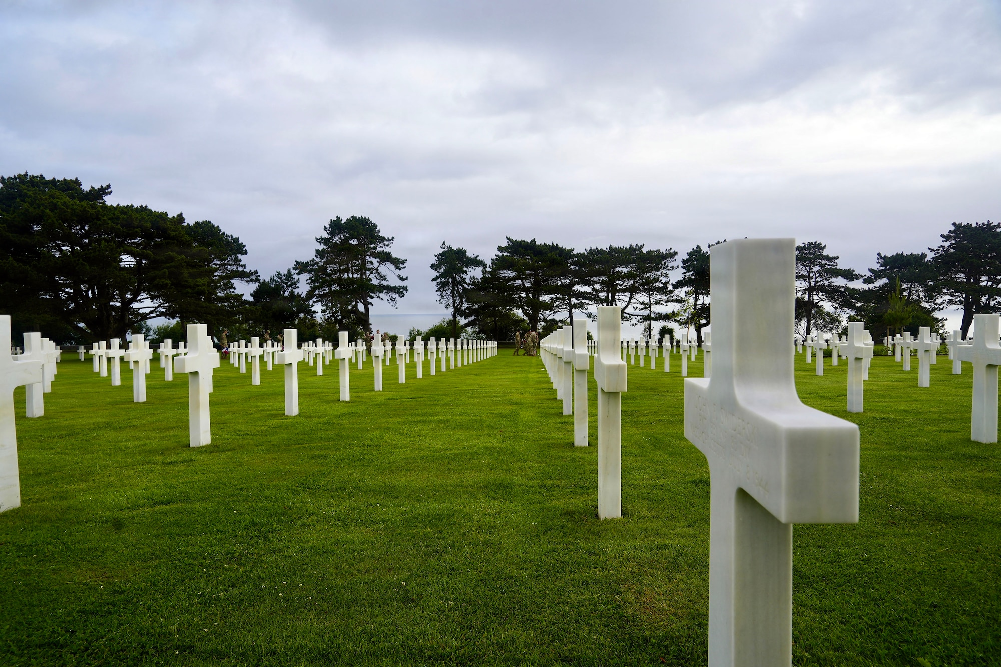 Rows of white headstones, mostly crosses, line a green field with trees in the distrance.