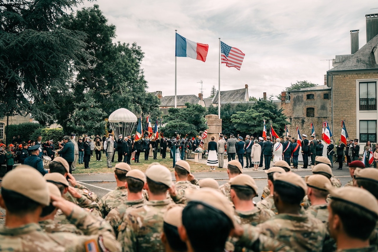 Soldiers stand in formation and salute amid a crowd standing outdoors by a French and a U.S. flag.